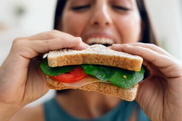 Beautiful sporty woman eating healthy sanwich while looking at camera in the kitchen at home