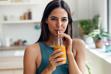 Beautiful sporty woman drinking healthy orange juice while standing in the kitchen at home