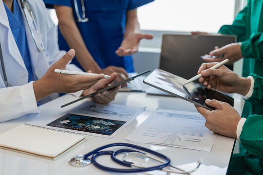 A multi-ethnic medical team is meeting with doctors in white lab coats and surgical gowns who sit at tables to discuss patient records, find a cure. Image of a group of doctors talking together