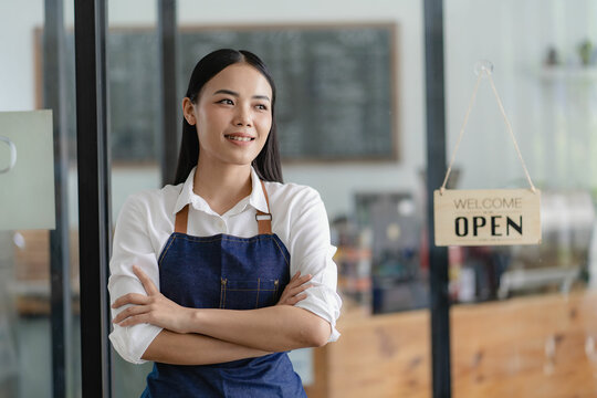 A Beautiful Asian Business Owner Stands At The Door Preparing To Take Orders From Customers. A Young Barista In An Apron Holds A Tablet And Stands In Front Of The Coffee Shop Door With An Open Sign.