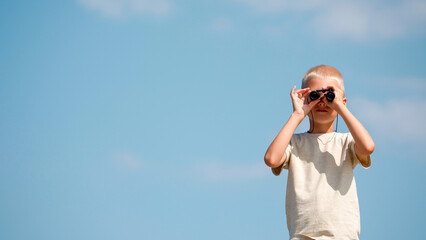 Portrait of a fair-haired European schoolboy looking through binoculars directly into the camera. An inquisitive child plays explorer or traveler, develops imagination. Active and healthy lifestyle.