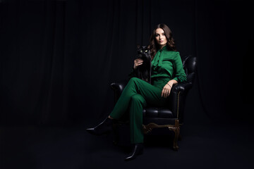 Fototapeta premium A young woman in a green suit holds a black cat in her hands while sitting in a chair.