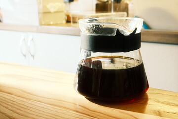 A freshly brewed black filter coffee in a shining glass decanter sitting on a wooden tabletop lighted with sunrays