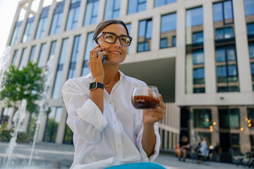 Fototapeta premium Positive female freelancer drinking coffee and talking phone sitting on modern building background 