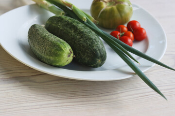 White Plate with fresh vegetables on wooden table. Two cucumbers, red cherry tomatoes, green cracked tomato with vertical splits, green Chives.  Healthy food, side view, copy space