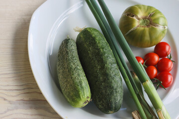 Set of fresh vegetables on the white plate. Two cucumbers, red cherry tomatoes, green cracked tomato with vertical splits, green Chives. Plate with raw food. Healthy food concept. Flat lay. Top view