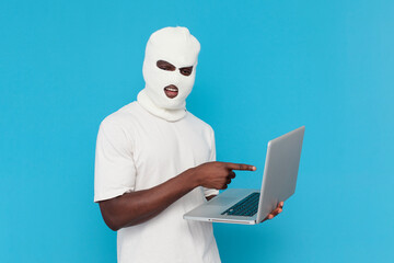 african american male hacker in white balaclava uses laptop on blue isolated background, bully in...