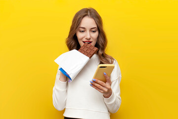 young girl eats big chocolate bar and uses smartphone and on yellow isolated background, woman with sweet food is typing