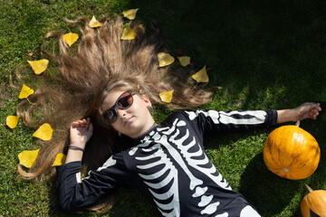 girl 10 - 12 years old in a black skeleton costume celebrates Halloween outdoors, lies on the...