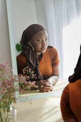 Pensive ethnic female in hijab looking in mirror