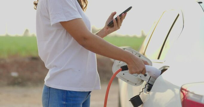 World Environment Day. save world save life. Hispanic female driver electric car holding electrical plug connector to charge the batteries hybrid car at home or charging station. Using smartphone