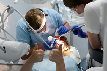 professional teeth cleaning in a dental clinic, removing stones and plaque, young specialists working with a woman, cleaning a saliva ejector, sucking out saliva, applying paint to the teeth - obrazy, fototapety, plakaty