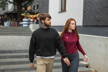 Beautiful fashion young couple in urban clothes with hoodies walking together hand in hand in the city. Handsome brutal young hipster man with a beard with hairstyle and a pretty redhead woman