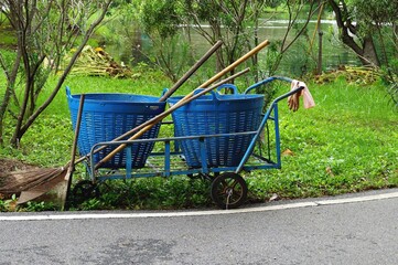 A cleaning cart with a blue plastic basket and  brooms stay beside a street in the park