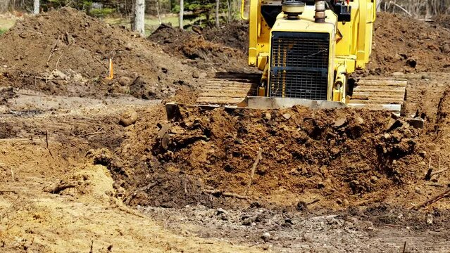 A yellow Bulldozer pushes brown soil toward camera as it levels the ground in preparation for the building of a new home.