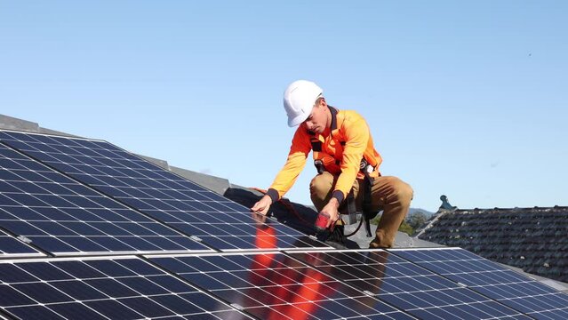 Solar panel technician with drill installing solar panels on house roof on a sunny day.