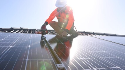 Solar panel technician with drill installing solar panels on house roof on a sunny day.