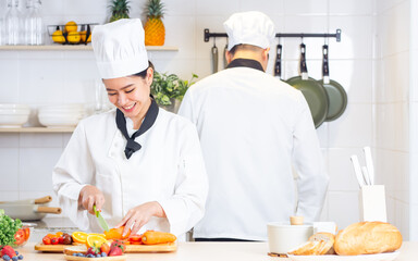 Two Asian professional couple chef wearing white uniform, hat, helping each other, preparing meal,cooking in kitchen together, smiling with happiness and confidence. Food Concept.