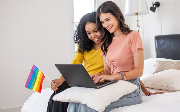 Portrait two diverse LGBT lesbian or female friends wearing casual clothes, sitting on bed in bedroom, using laptop, shopping online, smiling with happiness. Lifestyle Concept.