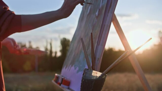 creative hobby, talented female artist draws picture with paints and brushes using an easel in park backdrop of sunlight, close-up
