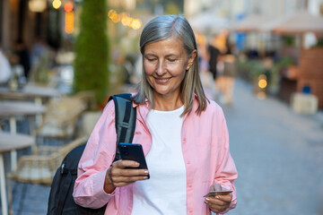 Senior mature female tourist with backpack and phone walking in evening city, gray-haired retired...