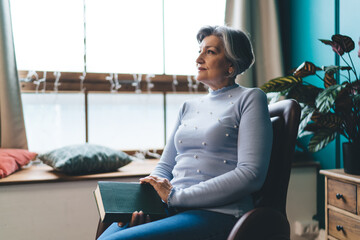 Thoughtful mature woman sitting on armchair with book
