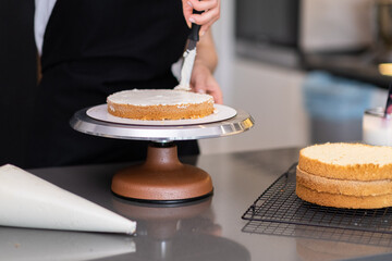 Female confectioner smearing cream around fresh cake layers with knife standing at kitchen table...
