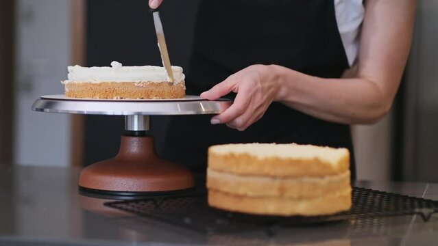 Female confectioner smearing cream around fresh cake layers with knife standing at kitchen table woman in apron making organic gluten free pastry for events at home
