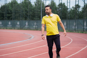 Bearded man in yellow t-shirt with wireless headphones standing on empty running track with red surface and white marking at contemporary sports ground in urban park