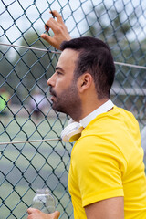 Interested sports fan wearing earphones eagerly observes the training session of a local soccer team on the urban arena, glimpsing through the metal fence grid. 