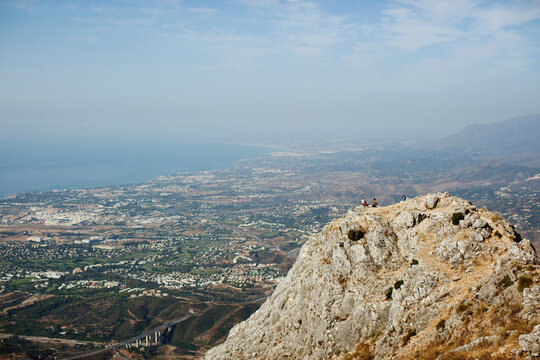 Two Mountain Climbers Sitting On A Peak Overlooking The City; Marbella, Malaga, Andalusia, Spain