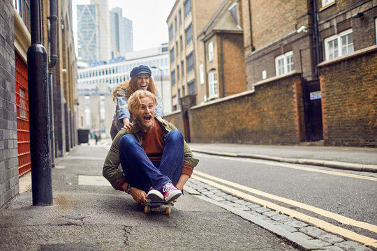 Happy young Caucasian redhead couple riding a skateboard while on a date in London UK