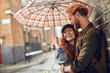 Young redhead couple using a umbrella to avoid the rain while on a date in London UK