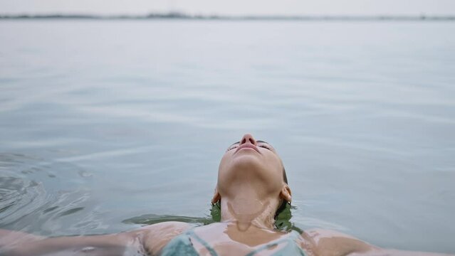 A young adult woman relaxes lying on the water surface in lake, eyes closed contentedly as she enjoys a dip in the water. woman standing up from water and looking at camera.
