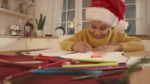 Stab shot of pretty 6 year old African American girl in Santa hat sitting at kitchen table and drawing pictures on Christmas holidays