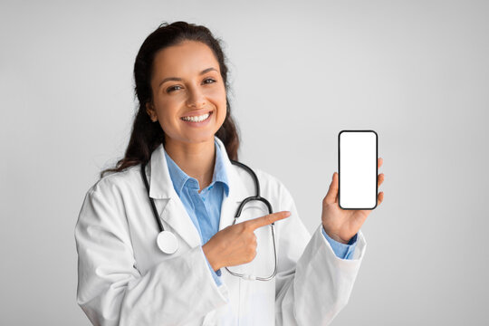 Smiling female physician showing and pointing at blank cellphone screen, standing on light wall background, mockup