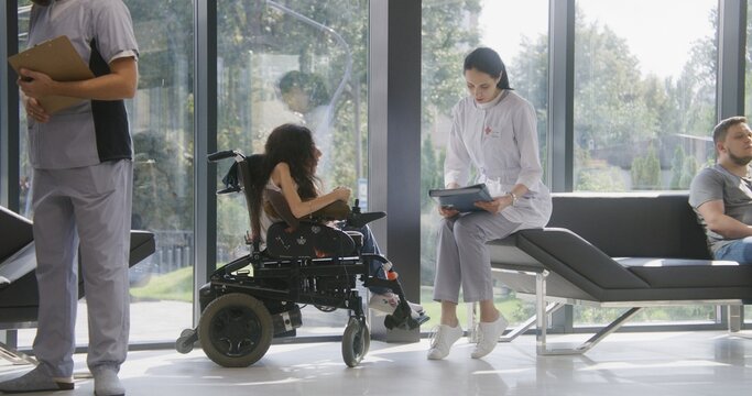 Woman in wheelchair talks to doctor in hospital or clinic lobby. Female physician sits on couch, consults patient with physical disability, uses mobile phone. Waiting area of modern medical facility.