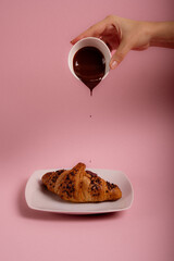 Chocolate croissant on the white plate.hand pouring chocolate onto a croissant. Pink background. 
