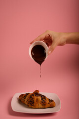 Chocolate croissant on the white plate.hand pouring chocolate onto a croissant. Pink background. 