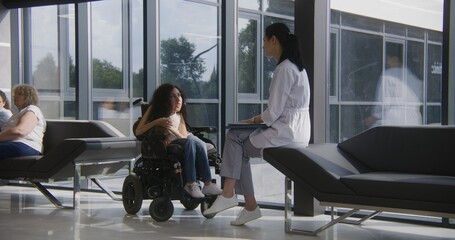 Woman with disability in motorized wheelchair talks to doctor in modern hospital or clinic lobby....