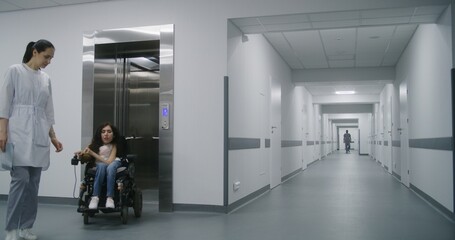 Woman in motorized wheelchair leaves elevator with doctor, rides along clinic corridor. Female...