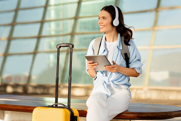 Smiling young lady in wireless headphones with suitcase typing on tablet, waiting for transport,...