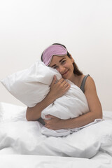 Morning bliss: a beautiful lady, wearing a sleep mask, smiles, clutching a pristine white pillow on her bedroom's comfort. 