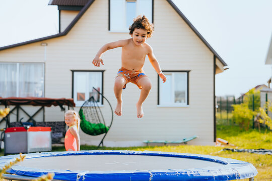Cheerful caucasian little boy jumping on trampoline against house and meadow. Active Spanish kid playing at country house yard. Childhood, freedom, entertainment. - Powered by Adobe