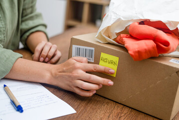 Woman pastes inscription Return on yellow sticker on cardboard box on wooden desk in premise bright things lie on top of parcel for return to online store