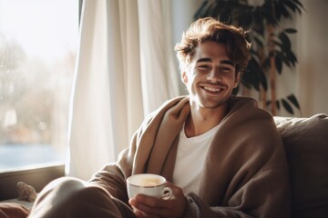 Cheerful young man sipping his coffee while wrapped in a warm blanket on the couch at home. The room emanates a cozy autumn-winter atmosphere, with soft lighting casting a gentle glow. Generative ai. - obrazy, fototapety, plakaty