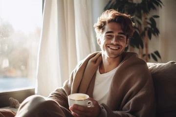 Schilderijen op glas Cheerful young man sipping his coffee while wrapped in a warm blanket on the couch at home. The room emanates a cozy autumn-winter atmosphere, with soft lighting casting a gentle glow. Generative ai. © StockWorld
