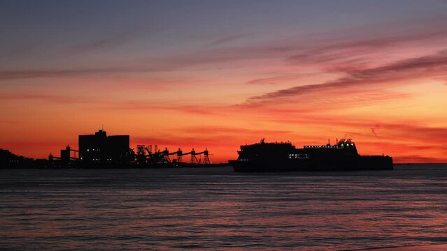 Passenger and vehicle ferry ship vessel silhouette in sea against dramatic sunset sky with Trafaria port terminal background in Lisbon, Portugal