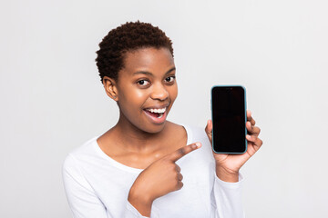 Adorable excited afro american woman holding cell phone smertphone in hands pointing at empty black screen standing on white background in studio mobile app concept banner advertisement - obrazy, fototapety, plakaty
