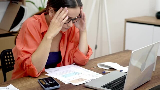Financial Confusion: In glasses, a stressed woman at a table looks at paper bills, perplexed by the high payment rates. 